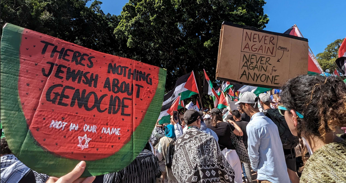 Protest scene with a watermelon sign reading 'there's nothing Jewish about genocide, not in our name' and 'never again means never again for anyone'