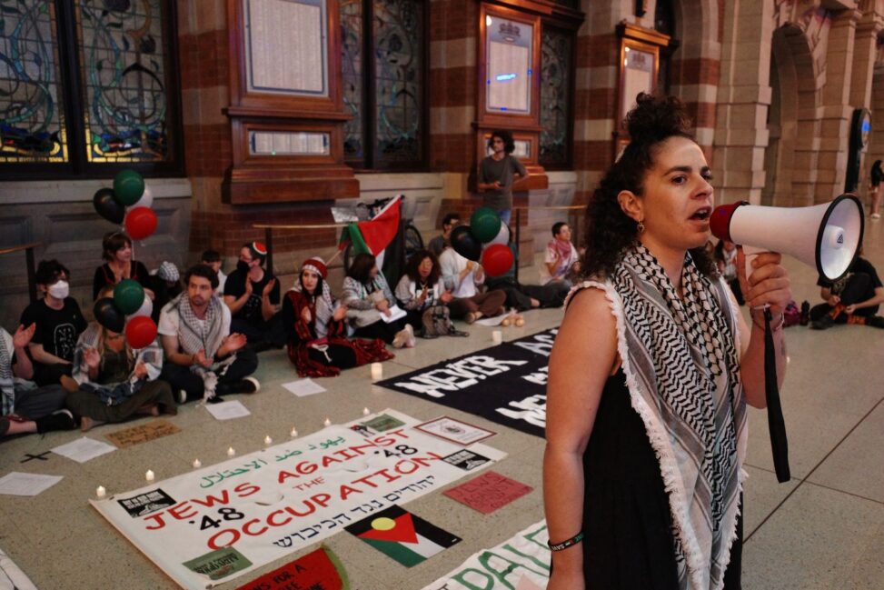 Protesters sit on the ground, around banners which state Jewish support Palestinian liberation and Palestinian flags. One woman stands at the front, leading a chant. She is wearing a keffiyeh and holding a megaphone.